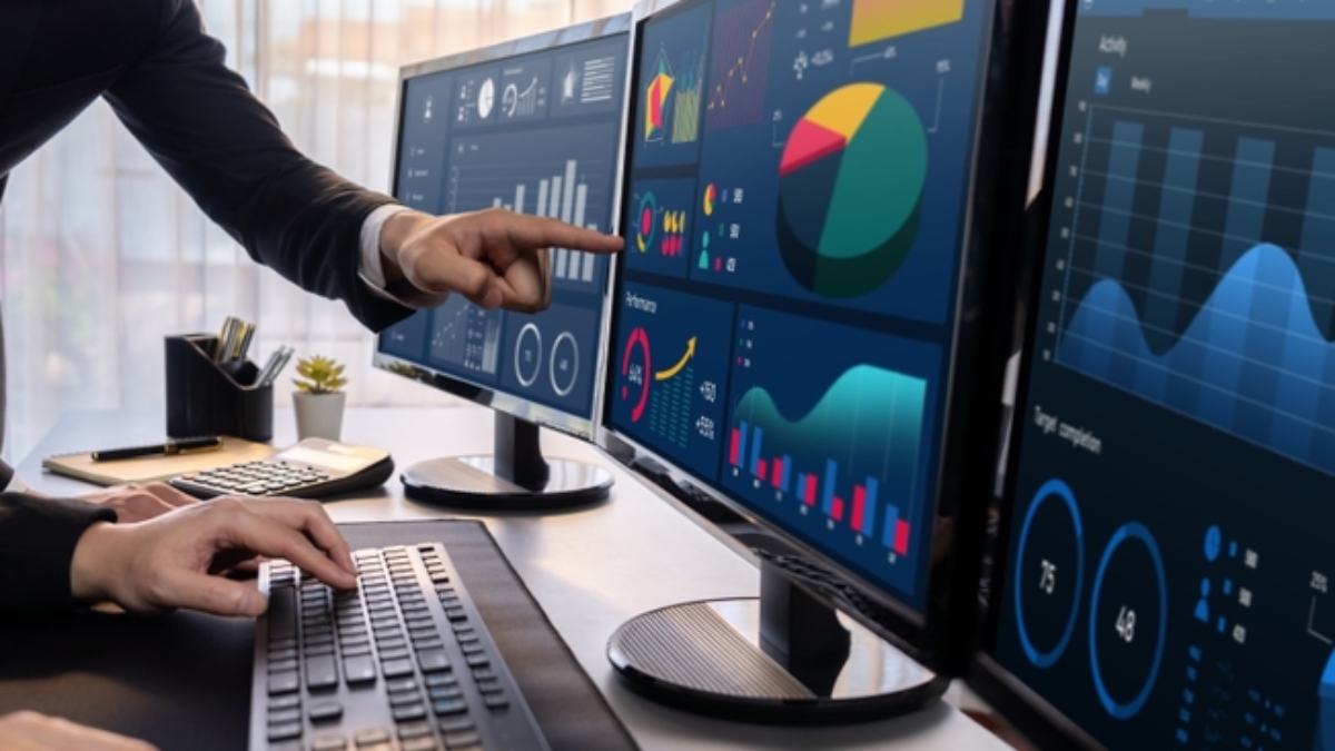 Two people analyze colorful financial charts and data visualizations on multiple monitors in an office, utilizing Microsoft Business Solutions—one points at a screen while the other types on a keyboard.