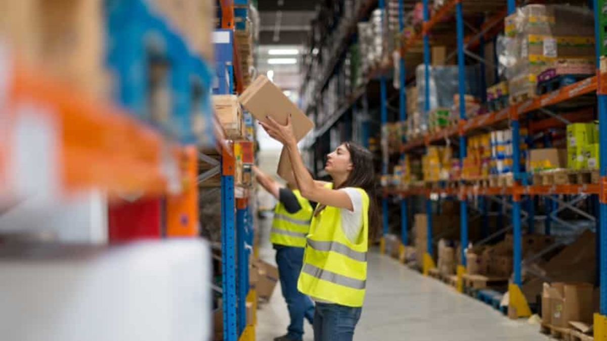 A woman in a yellow safety vest places a box on a high shelf in a warehouse, while another worker in a vest organizes items on a nearby shelf. Shelves are filled with various boxes and products.