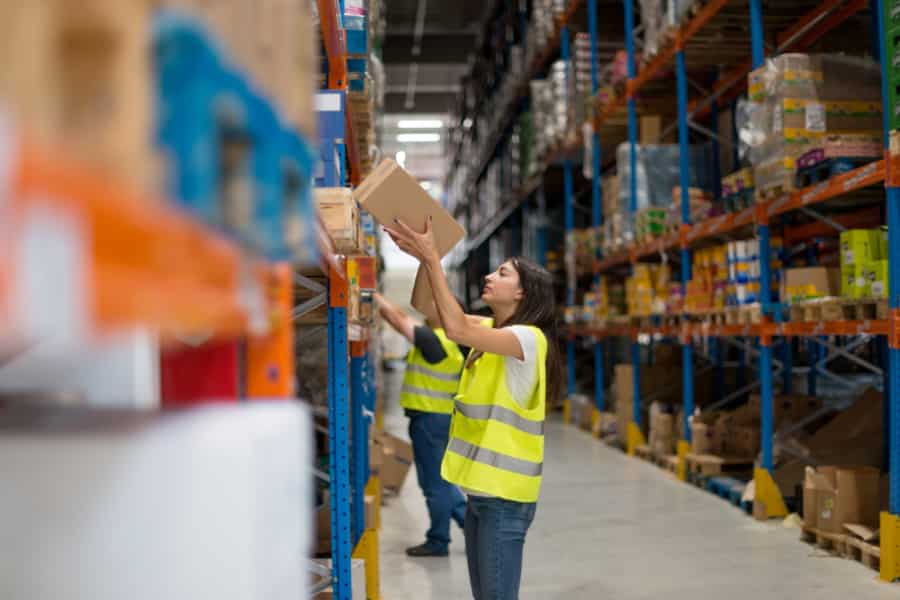 A woman in a yellow safety vest places a box on a high shelf in a warehouse, while another worker in a vest organizes items on a nearby shelf. Shelves are filled with various boxes and products.