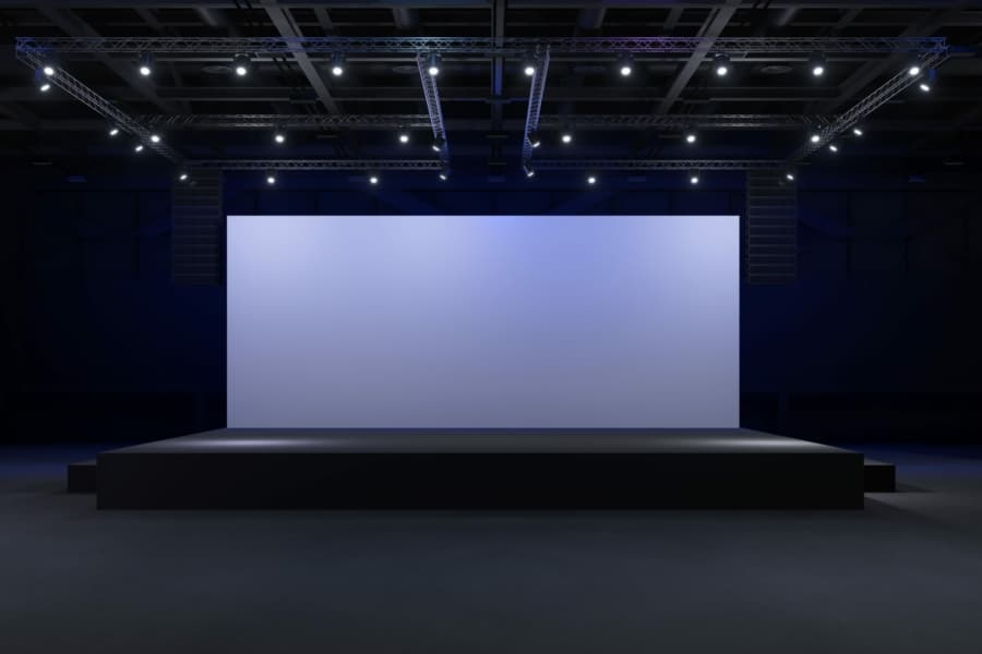 A large, empty stage with a blank screen is illuminated by spotlights in a dark auditorium, setting the scene for a WMS Implementation Case Study presentation or performance.