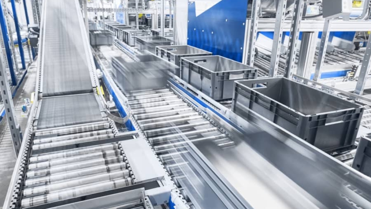 A modern automated conveyor system transports black plastic bins along metal rollers in a bright, industrial warehouse setting. The machinery and containers are in motion, suggesting efficient, high-speed operations.