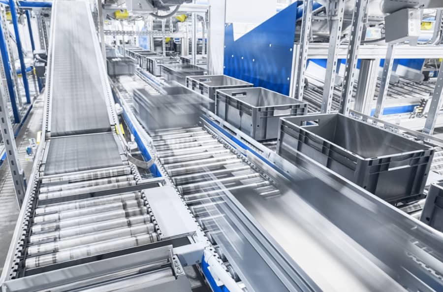 A modern automated conveyor system transports black plastic bins along metal rollers in a bright, industrial warehouse setting. The machinery and containers are in motion, suggesting efficient, high-speed operations.