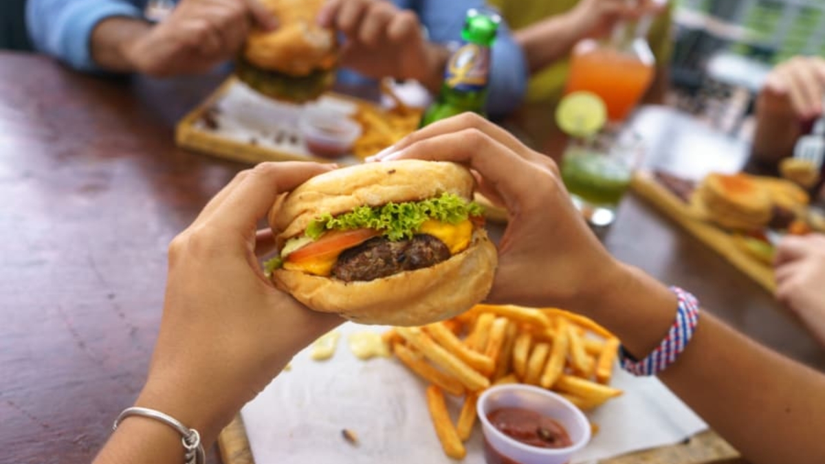 A person holds a cheeseburger with lettuce and tomato over a tray of fries and ketchup, with other people eating burgers and drinking colorful beverages at a wooden table.