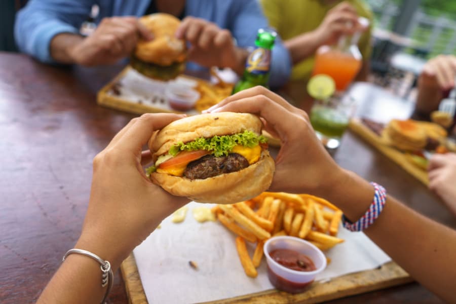 A person holds a cheeseburger with lettuce and tomato over a tray of fries and ketchup, with other people eating burgers and drinking colorful beverages at a wooden table.