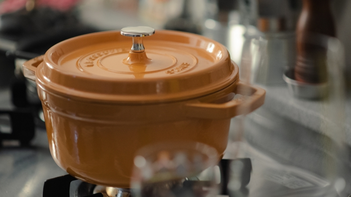 An orange dutch oven with a silver knob sits on a gas stovetop in a kitchen, with blurred kitchen items and utensils visible in the background.