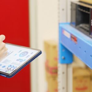 Person in blue uniform holds a tablet displaying WMS charts and graphs while standing near warehouse shelves with boxes.