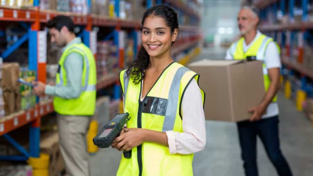 A smiling woman in a yellow safety vest holds a barcode scanner in a warehouse, with two coworkers working in the background among shelves stocked with boxes and goods.