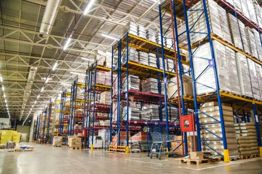 Large warehouse interior with tall blue and yellow racks stacked with pallets of boxes and goods. Wide aisles separate the shelves, and bright lighting illuminates the organized storage area.