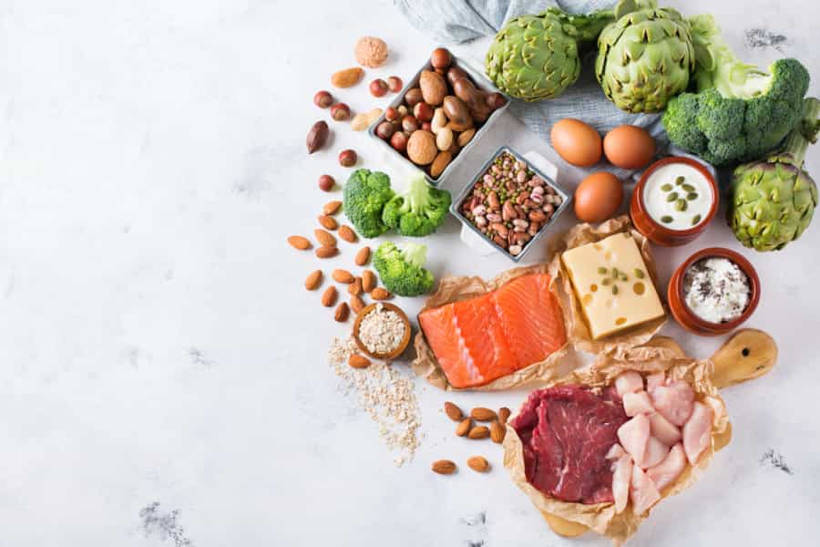 Assorted healthy foods including salmon, beef, chicken, cheese, eggs, nuts, seeds, yogurt, artichokes, broccoli, and beans arranged on a white surface.