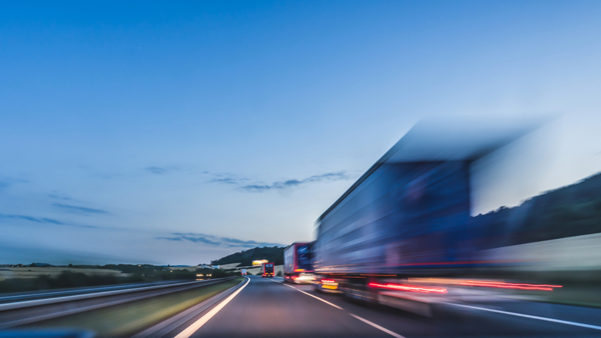 A blurred truck speeds down a highway at dusk, creating motion blur. The sky is clear with soft blue tones, and the distant landscape is faintly visible beside the road—capturing the essence of modern transportation sourcing in motion.
