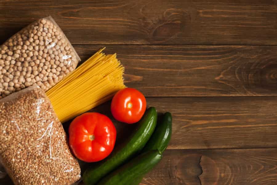 A flat lay of two tomatoes, two cucumbers, uncooked spaghetti, dried chickpeas, and grains on a dark wooden surface.