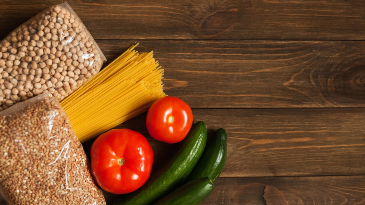 Chickpeas, spaghetti, and grains in plastic bags, along with two tomatoes and three cucumbers, are arranged on a wooden table. The food is placed in the lower left corner, leaving most of the table surface visible.