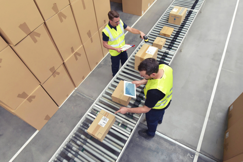 Two warehouse workers in yellow safety vests process parcels on a conveyor belt, scanning and checking boxes. Stacks of cardboard boxes are arranged along the sides of the conveyor in a large, organized warehouse space.