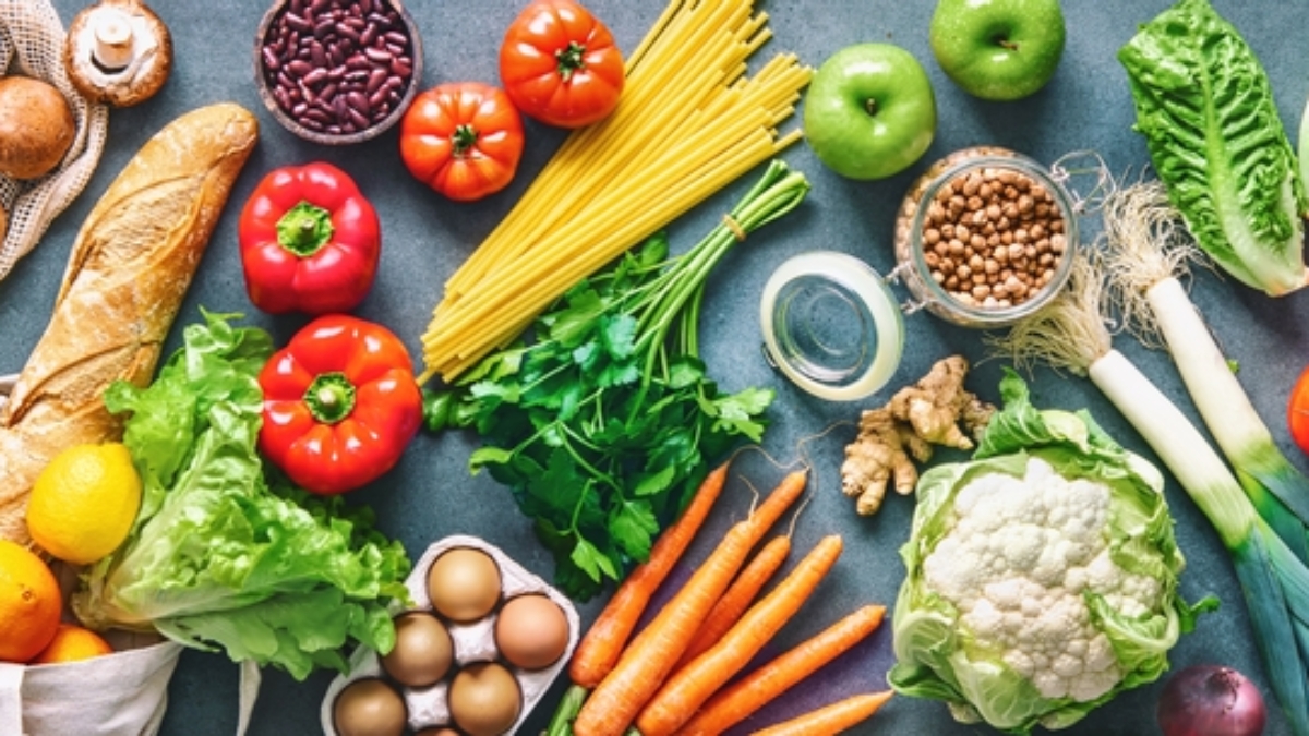 A variety of fresh foods including vegetables, fruits, bread, eggs, pasta, beans, olive oil, and milk are arranged on a table, showcasing healthy ingredients for balanced meals.