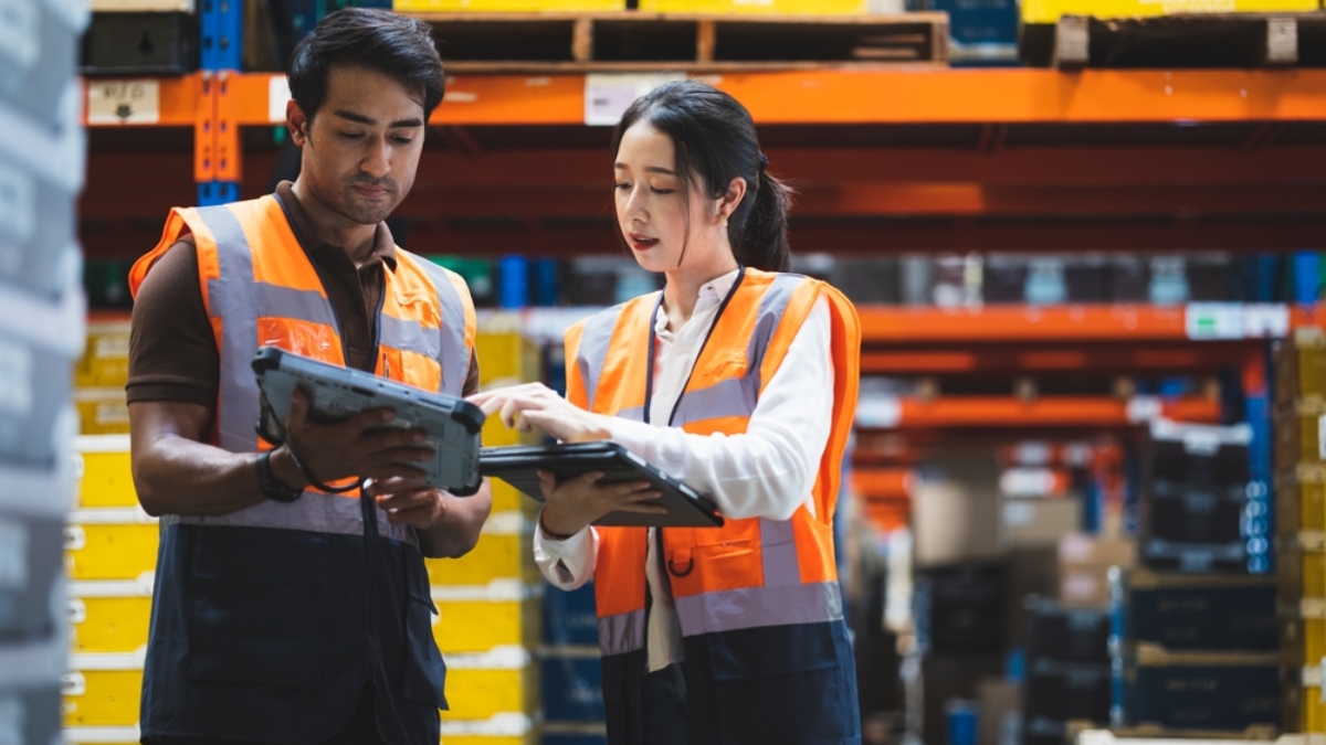 Two warehouse workers wearing orange safety vests review labor management information on tablets while standing in an aisle filled with shelves of yellow and blue storage bins.