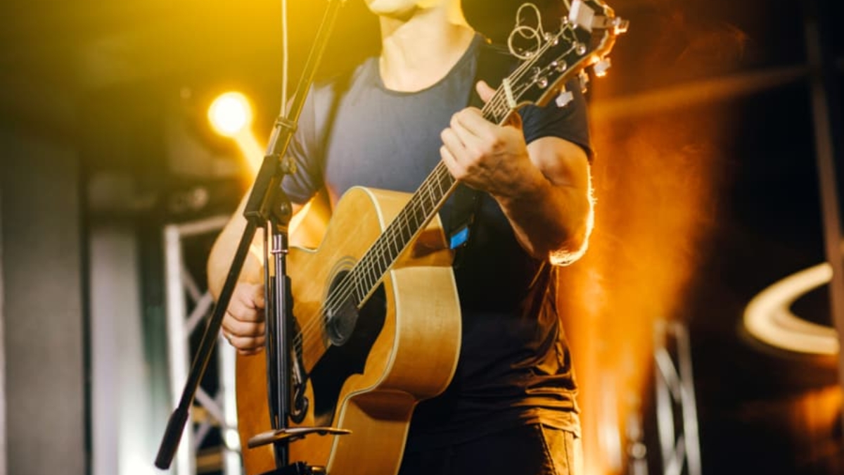 A person playing an acoustic guitar and singing into a microphone on stage, illuminated by bright yellow and orange stage lights.