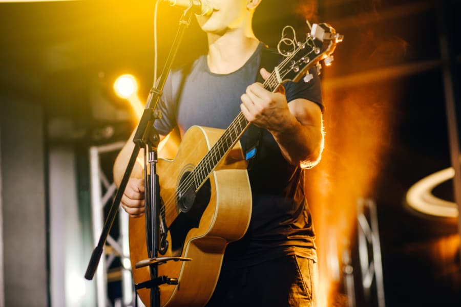 A person playing an acoustic guitar and singing into a microphone on stage, illuminated by bright yellow and orange stage lights.
