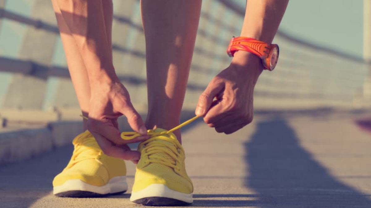 A person bends down to tie the yellow shoelaces on their bright yellow running shoes while standing on a sunlit outdoor pathway, wearing an orange watch on their wrist.