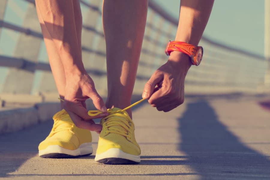 A person bends down to tie the yellow shoelaces on their bright yellow running shoes while standing on a sunlit outdoor pathway, wearing an orange watch on their wrist.