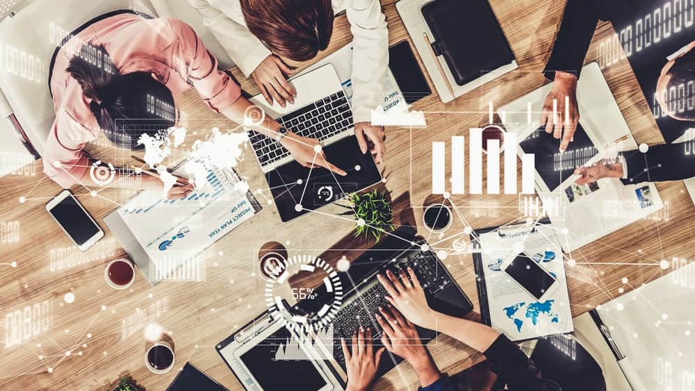A group of people work together at a table with laptops, tablets, documents, and coffee. Digital graphics overlay the scene, showing charts and data visualizations as they collaborate to select the right ERP system for their business needs.