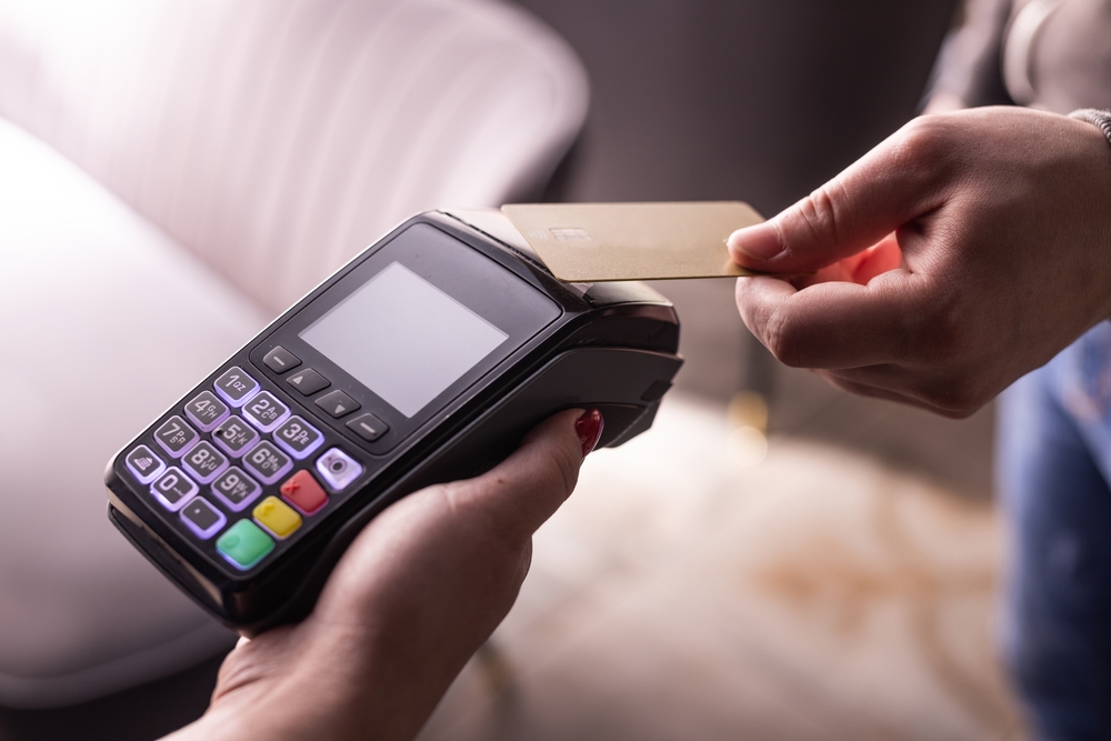 A person holds a payment terminal while another taps a gold credit card, showcasing how contactless payments can help automate everyday transactions.
