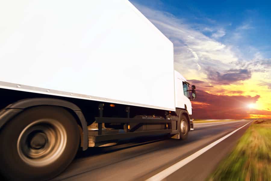 A white delivery truck speeds down a highway at sunset, framed by dramatic blue, orange, and yellow skies—a striking scene that captures the efficiency powered by modern transportation software.