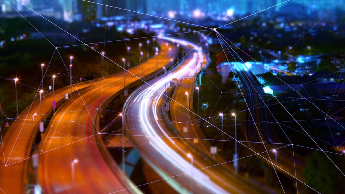 A city highway at night with blurred lights showing fast-moving traffic. Thin digital network lines are overlaid, symbolizing smart urban infrastructure and seamless connectivity—much like an OMS enhances what is supply chain strategy efficiency. Skyscrapers are visible in the background.
