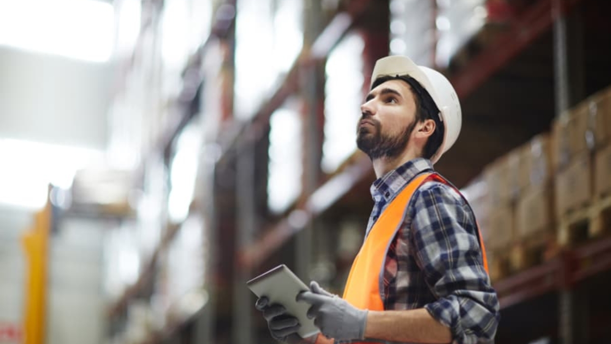 A warehouse worker wearing a hard hat, orange safety vest, gloves, and a plaid shirt uses a tablet for project management as he inspects shelves. The background shows stacked boxes on high racks.