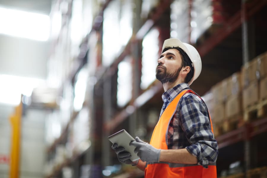A warehouse worker wearing a hard hat, orange safety vest, gloves, and a plaid shirt uses a tablet for project management as he inspects shelves. The background shows stacked boxes on high racks.