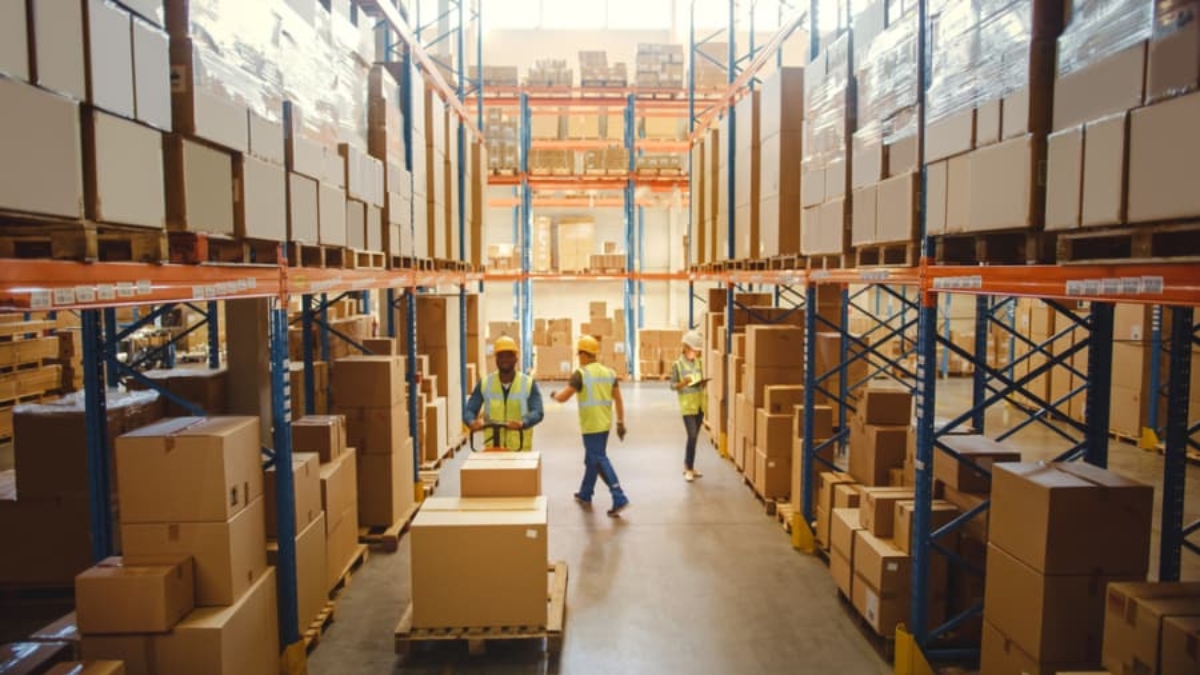 Warehouse workers in safety vests walk and talk between tall shelves filled with stacked boxes and packages during a Distribution Center Operational Assessment in a brightly lit warehouse.