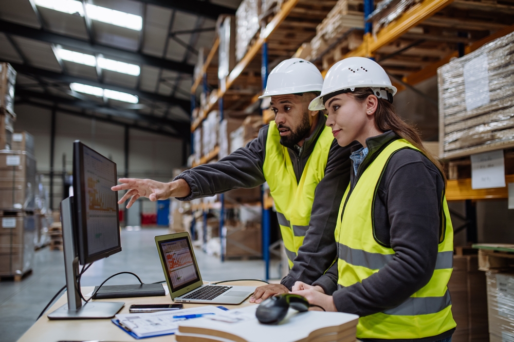 Warehouse,Manager,Talking,With,Logistics,Employee,In,Warehouse,,Planning,Transport Two warehouse workers in safety vests and hard hats review order accuracy on a computer monitor in a storage facility, surrounded by shelves with stacked boxes and paperwork on a desk.