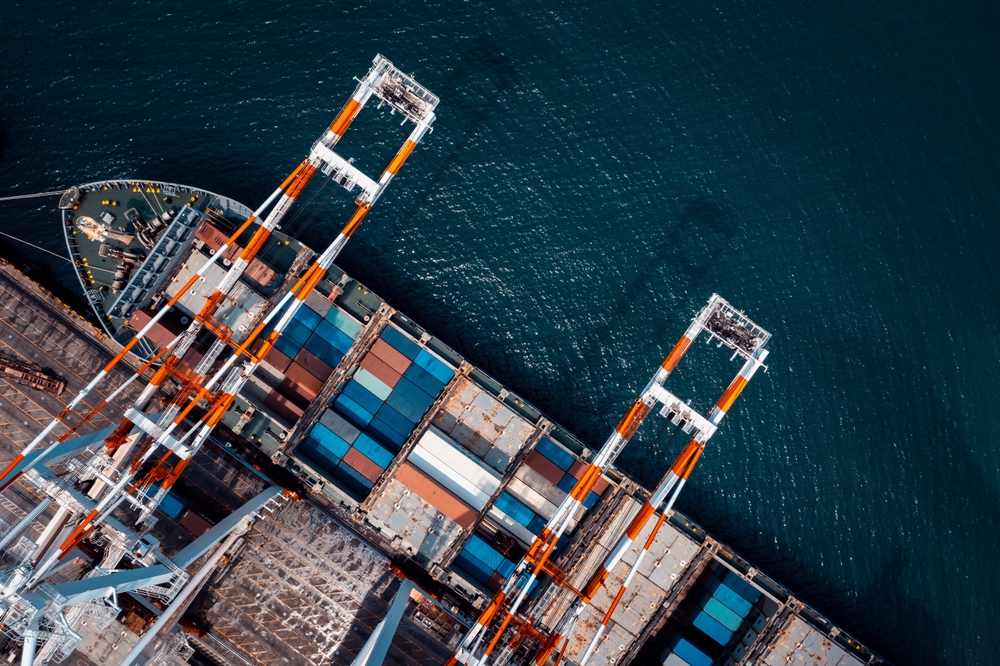 Aerial view of a cargo ship docked at a port, with colorful shipping containers and large orange-and-white cranes unloading goods—resembling the organized structure of Microsoft Fabric by the water.