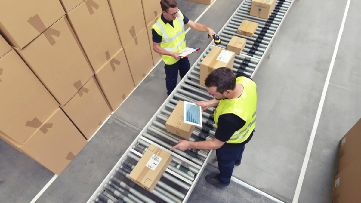 Two workforce members in yellow vests scan and check boxes on a conveyor belt in a warehouse. Stacks of cardboard boxes are visible nearby, and both workers are handling packages.
