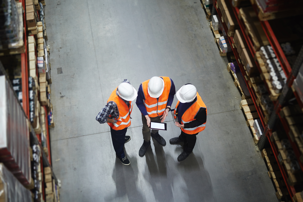 Three people in white hard hats and orange safety vests stand in a warehouse aisle, discussing what is supply chain strategy while looking at a tablet. Shelves filled with boxes line both sides of the concrete floor. View is from above.