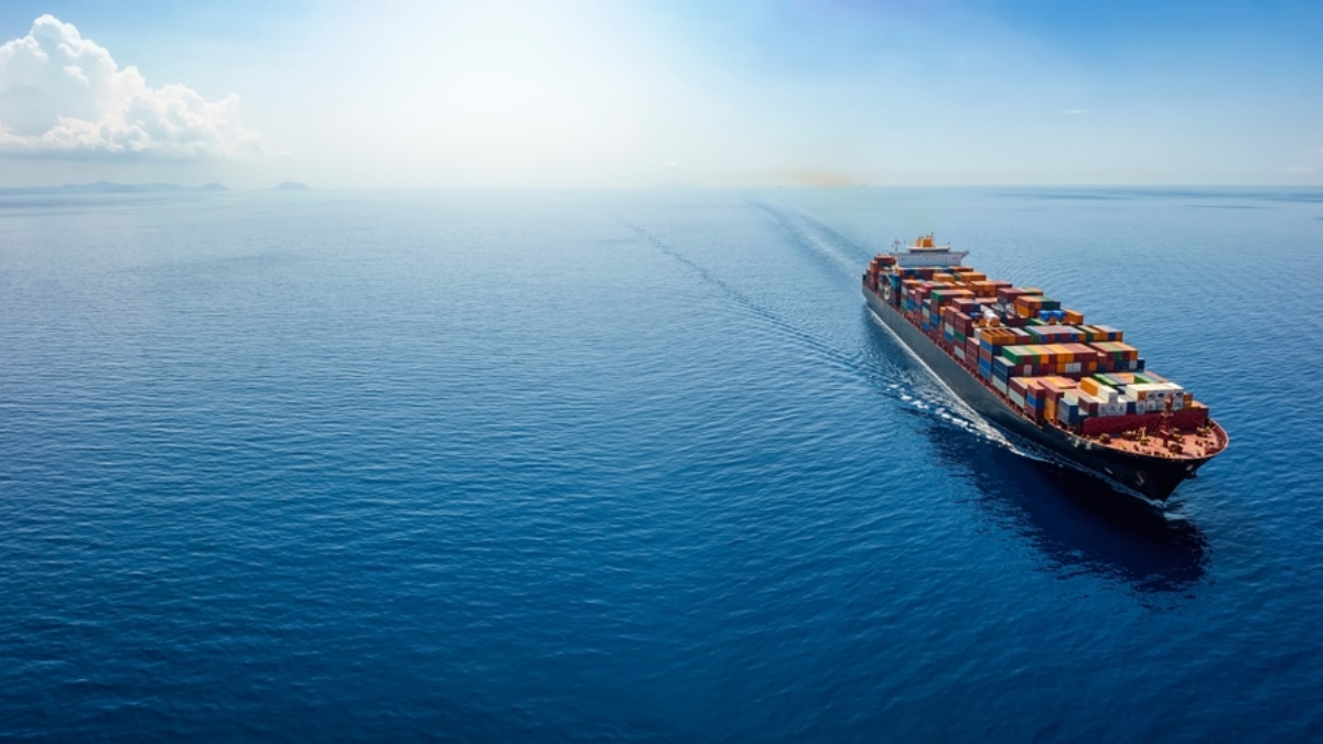 A large cargo ship loaded with colorful shipping containers sails across a calm blue ocean under a clear sky, leaving a wake behind.