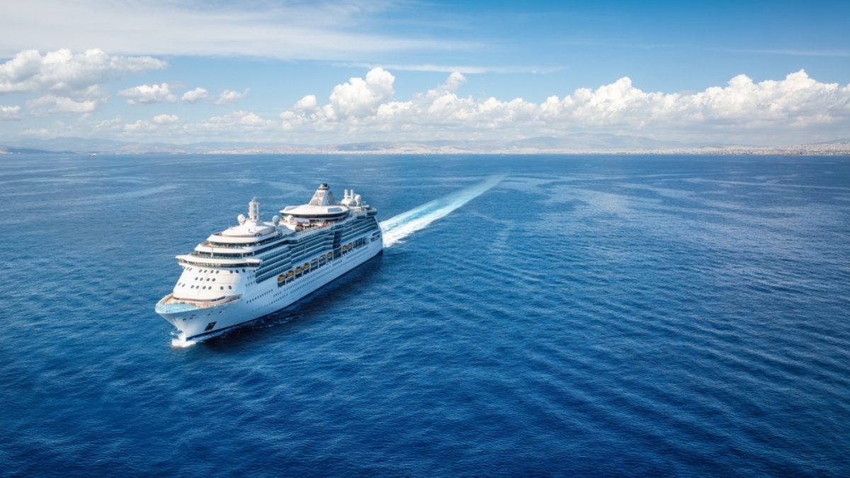 A large white cruise ship sails across a calm, deep blue sea under a bright sky with scattered clouds, leaving a wake behind it. Land is visible in the distance on the horizon.