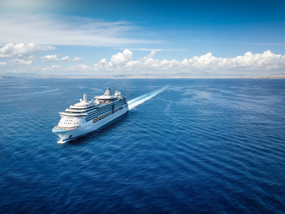A large white cruise ship sails across a calm, deep blue sea under a bright sky with scattered clouds, leaving a wake behind it. Land is visible in the distance on the horizon.