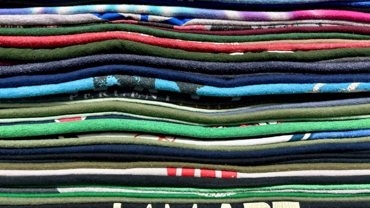 A close-up view of a neatly stacked pile of folded t-shirts in various colors and patterns, creating a layered, striped appearance.