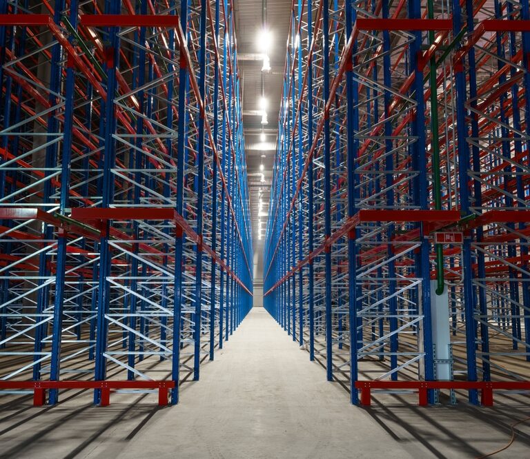 Empty metal storage racks extend in symmetrical rows inside a large warehouse, creating a sense of depth and perspective—an ideal setting for ai-powered inventory systems. Bright lights illuminate the clean concrete floor and steel framework.