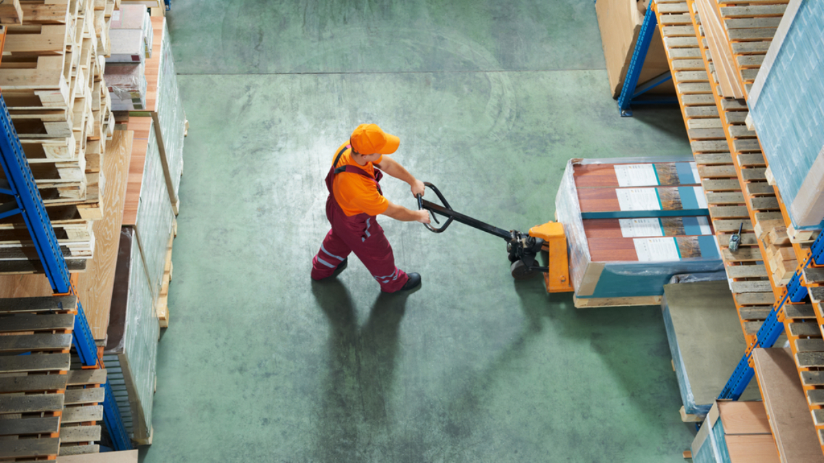 A workforce member in an orange shirt and cap uses a pallet jack to move boxed goods between shelves on a green concrete floor, viewed from above.