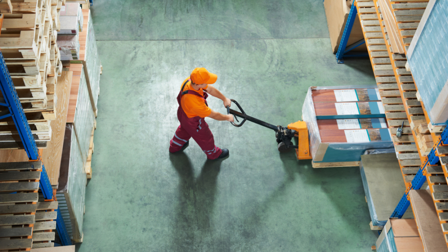 A workforce member in an orange shirt and cap uses a pallet jack to move boxed goods between shelves on a green concrete floor, viewed from above.
