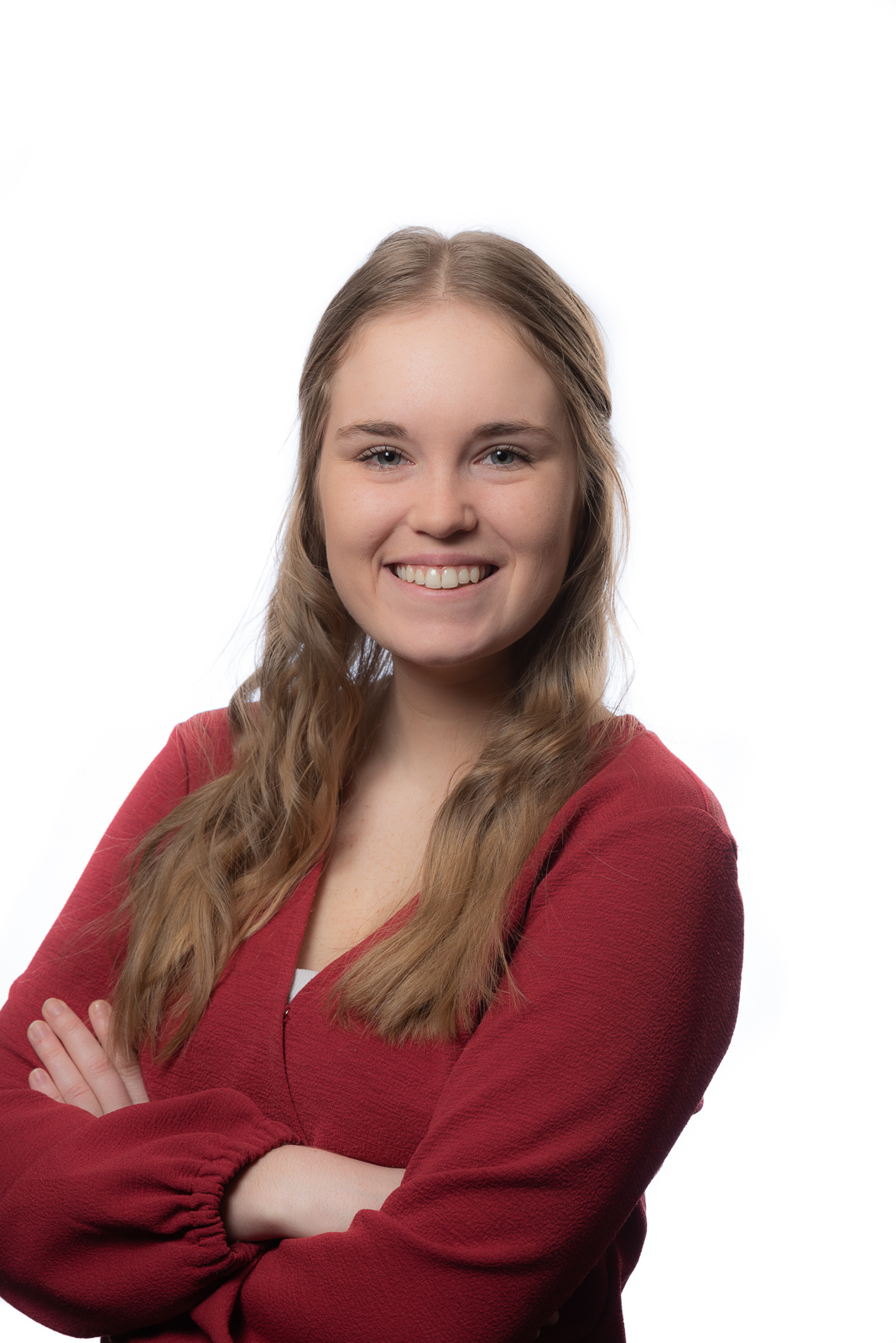 A young woman with long, light brown hair smiles confidently with her arms crossed, embodying the professionalism sought at enVista Careers. She is wearing a red top and stands against a plain white background.