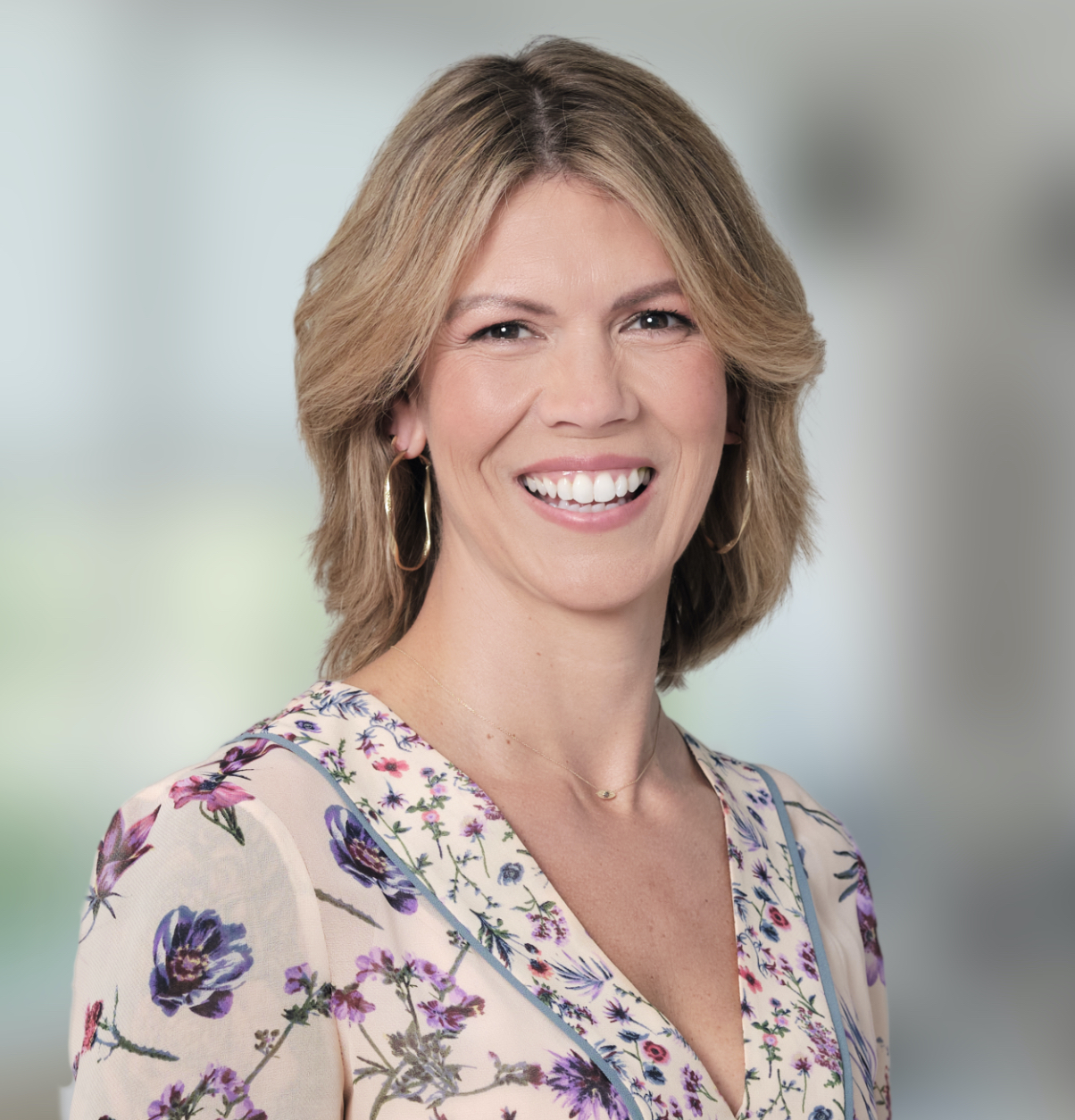 A smiling woman with shoulder-length blonde hair, wearing a floral blouse and hoop earrings, stands in front of a softly blurred indoor background—an inviting image for enVista Careers.