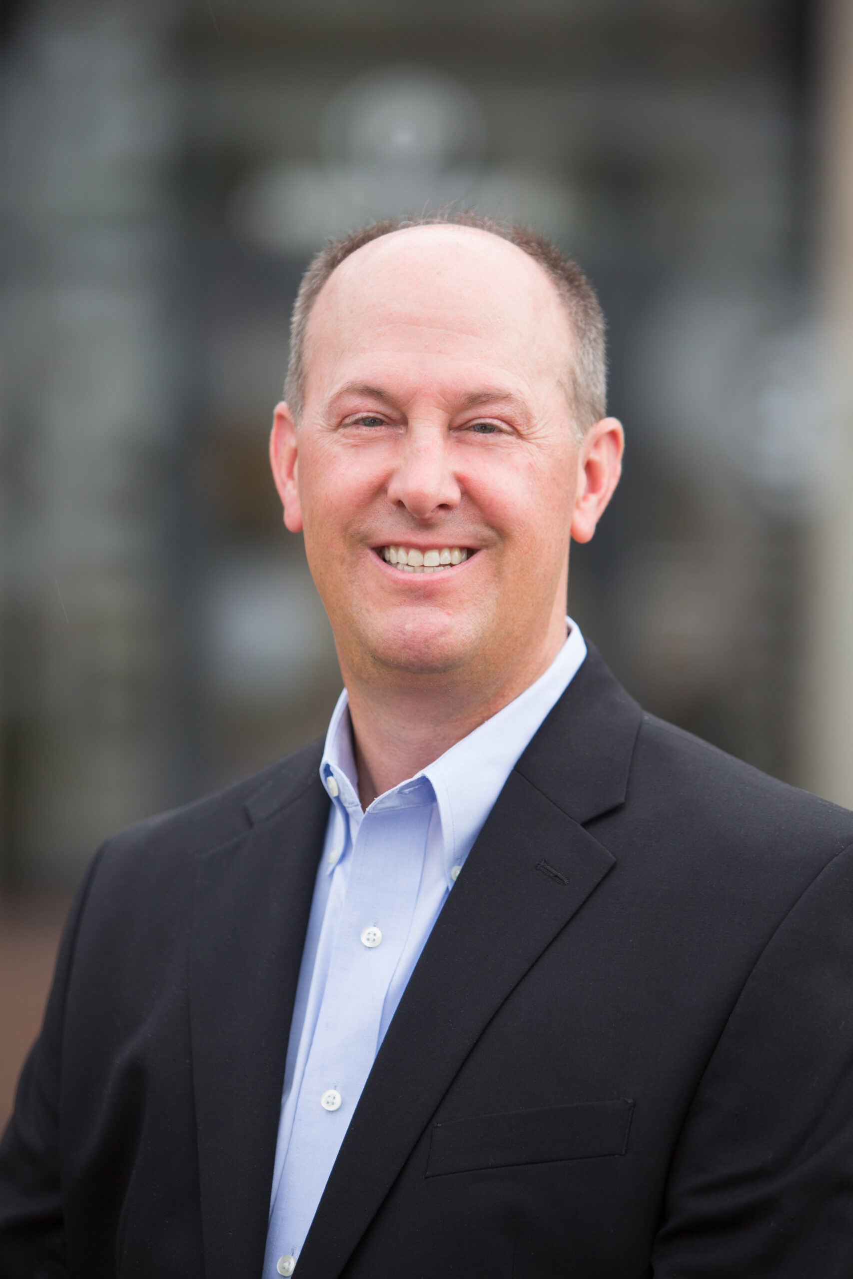 A man with short hair wearing a black suit jacket and light blue dress shirt smiles while standing outdoors, representing enVista Careers in front of a blurred background.