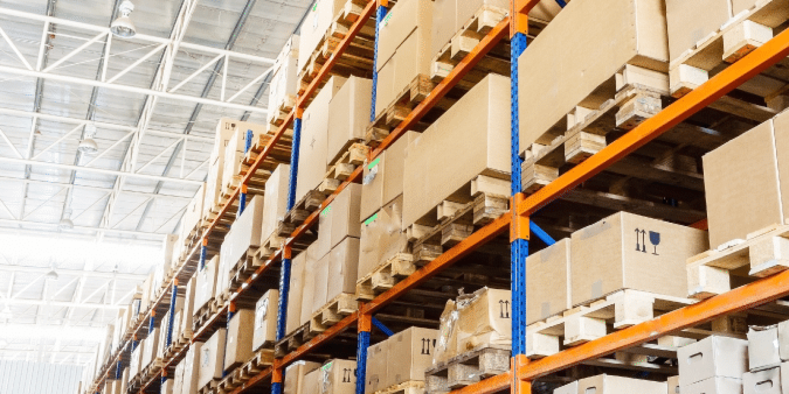 Large warehouse interior with tall blue and orange metal shelves stacked with brown cardboard boxes and wooden pallets, extending into the distance under a high ceiling with exposed beams.