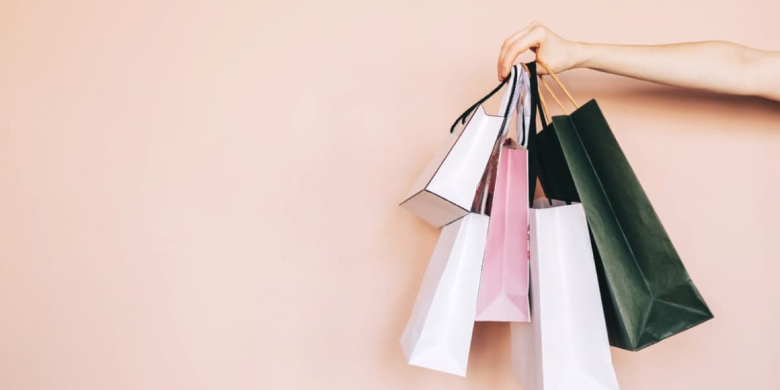 A hand is holding several colorful shopping bags by their handles against a light peach background, highlighting the variety that WMS can help organize. The bags vary in size and color, including white, pink, black, and dark green.