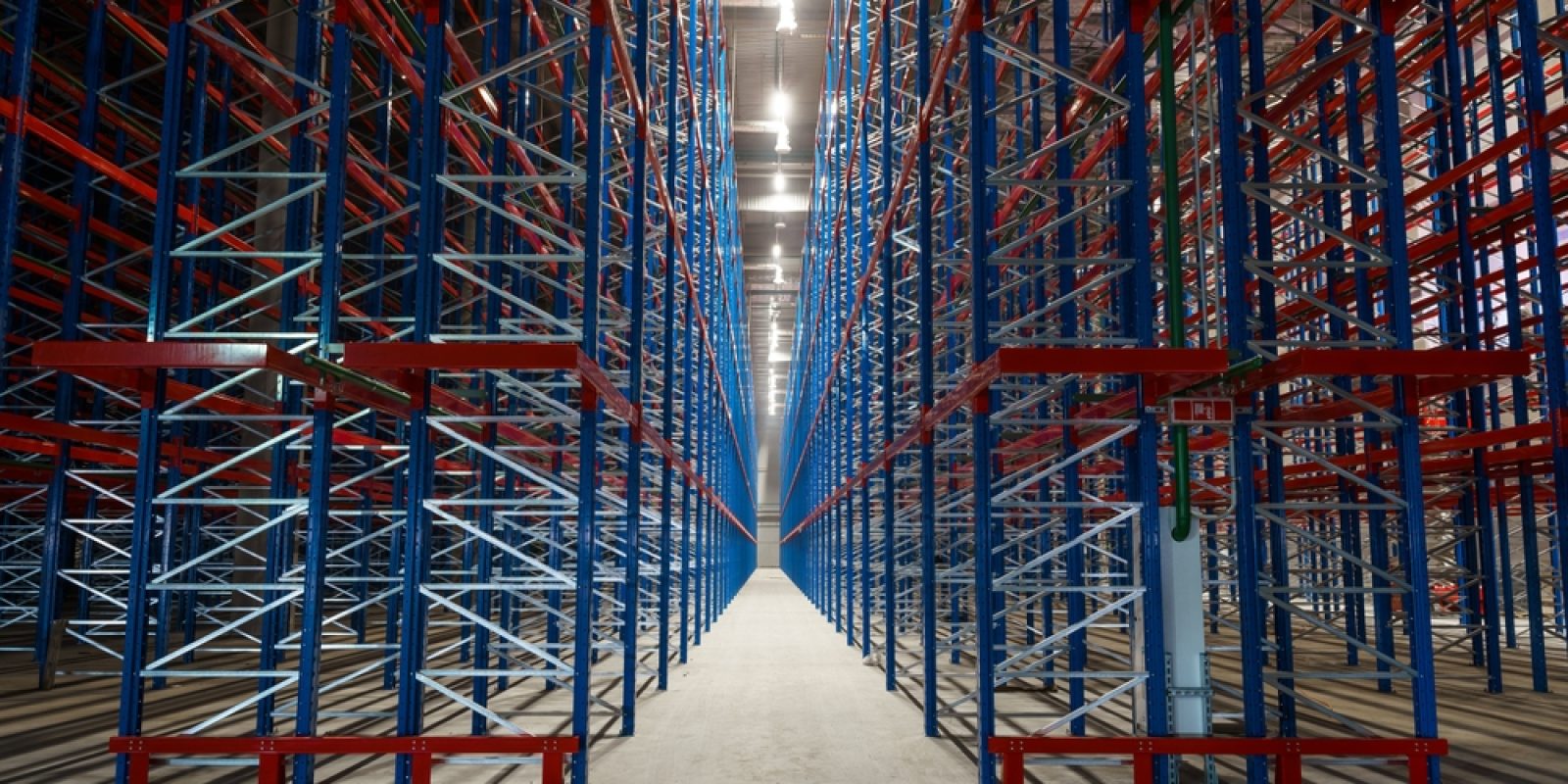 Empty metal storage racks extend in symmetrical rows inside a large warehouse, creating a sense of depth and perspective—an ideal setting for ai-powered inventory systems. Bright lights illuminate the clean concrete floor and steel framework.