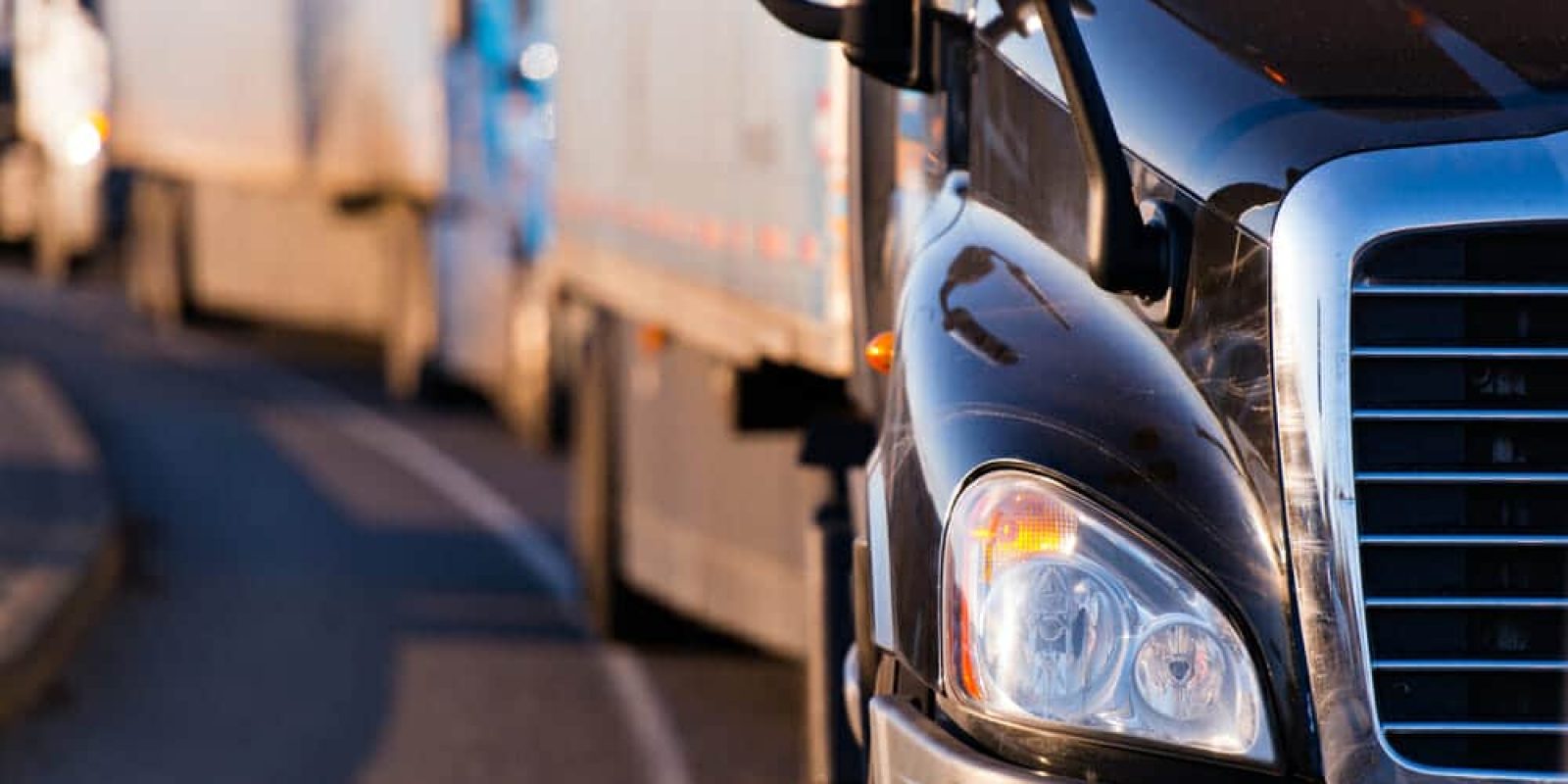 Close-up of the front of a black semi-truck driving on a road, with several other trucks visible in a line behind it, curving along the road.