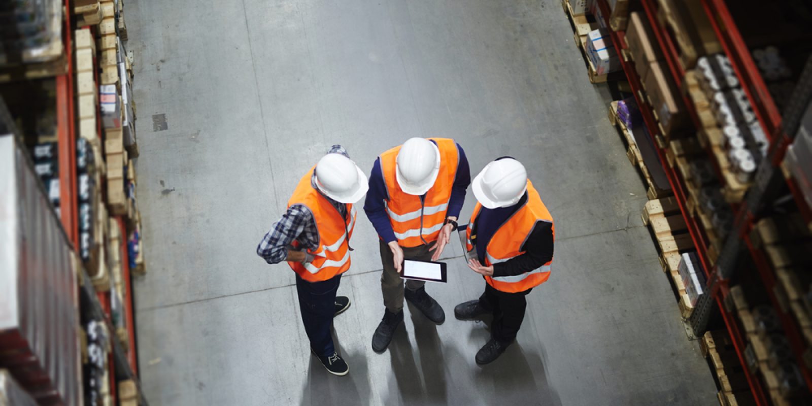 Three people in white hard hats and orange safety vests stand in a warehouse aisle, discussing what is supply chain strategy while looking at a tablet. Shelves filled with boxes line both sides of the concrete floor. View is from above.